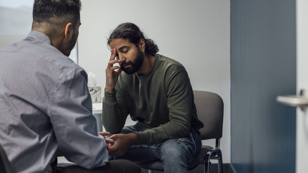 Over-the-shoulder shot of a doctor and man. The man looks sad and depressed after hearing news from the doctor. The man's eyes are closed and has one hand covering part of his face.
