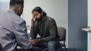 Over-the-shoulder shot of a doctor and man. The man looks sad and depressed after hearing news from the doctor. The man&#039;s eyes are closed and has one hand covering part of his face.