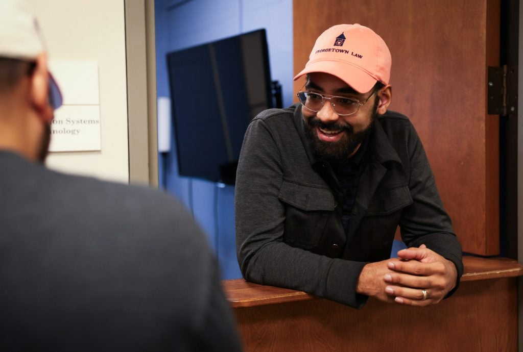A man wearing an orange Georgetown Law baseball cap leans out of the Information Systems Technology service desk and smiles