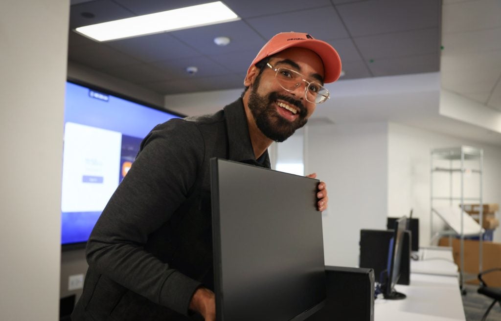 A man in an orange baseball cap smiles as he installs a laptop in a classroom