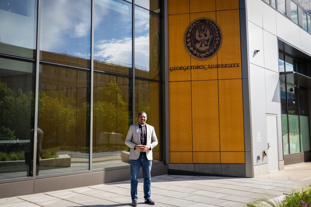 A man in a clerical collar stands outside a glass building with Georgetown's seal on the outside 