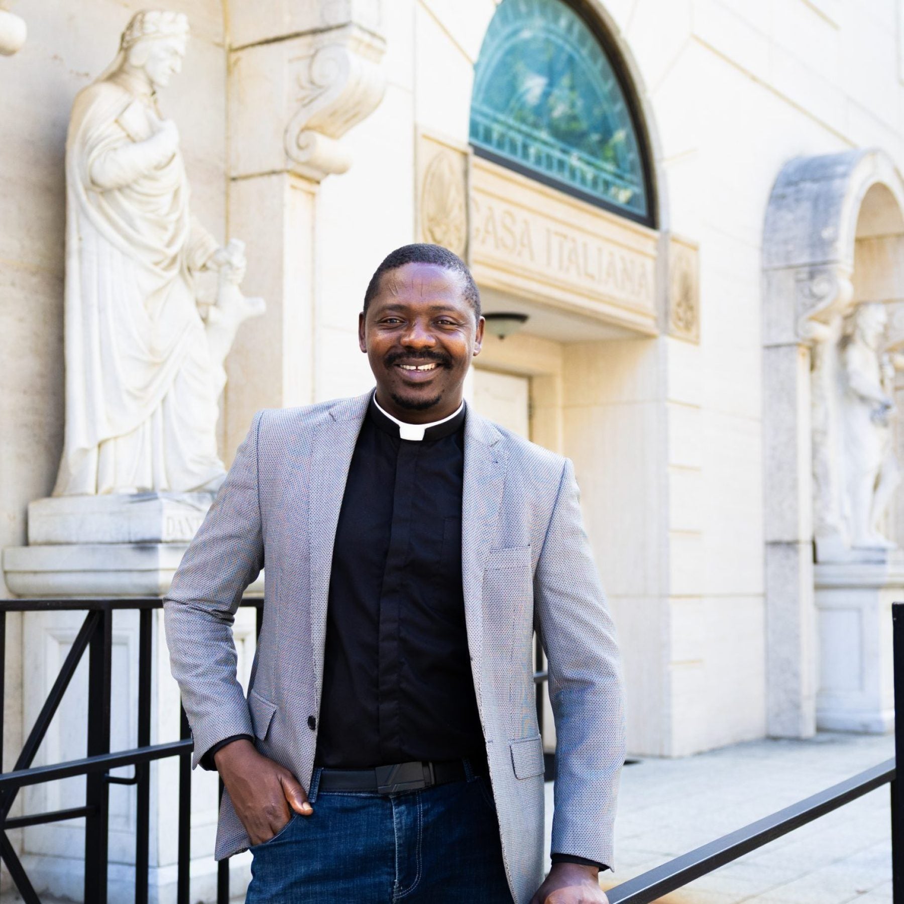 A man wearing a priest&#039;s collar and gray blazer smiles in front of a church