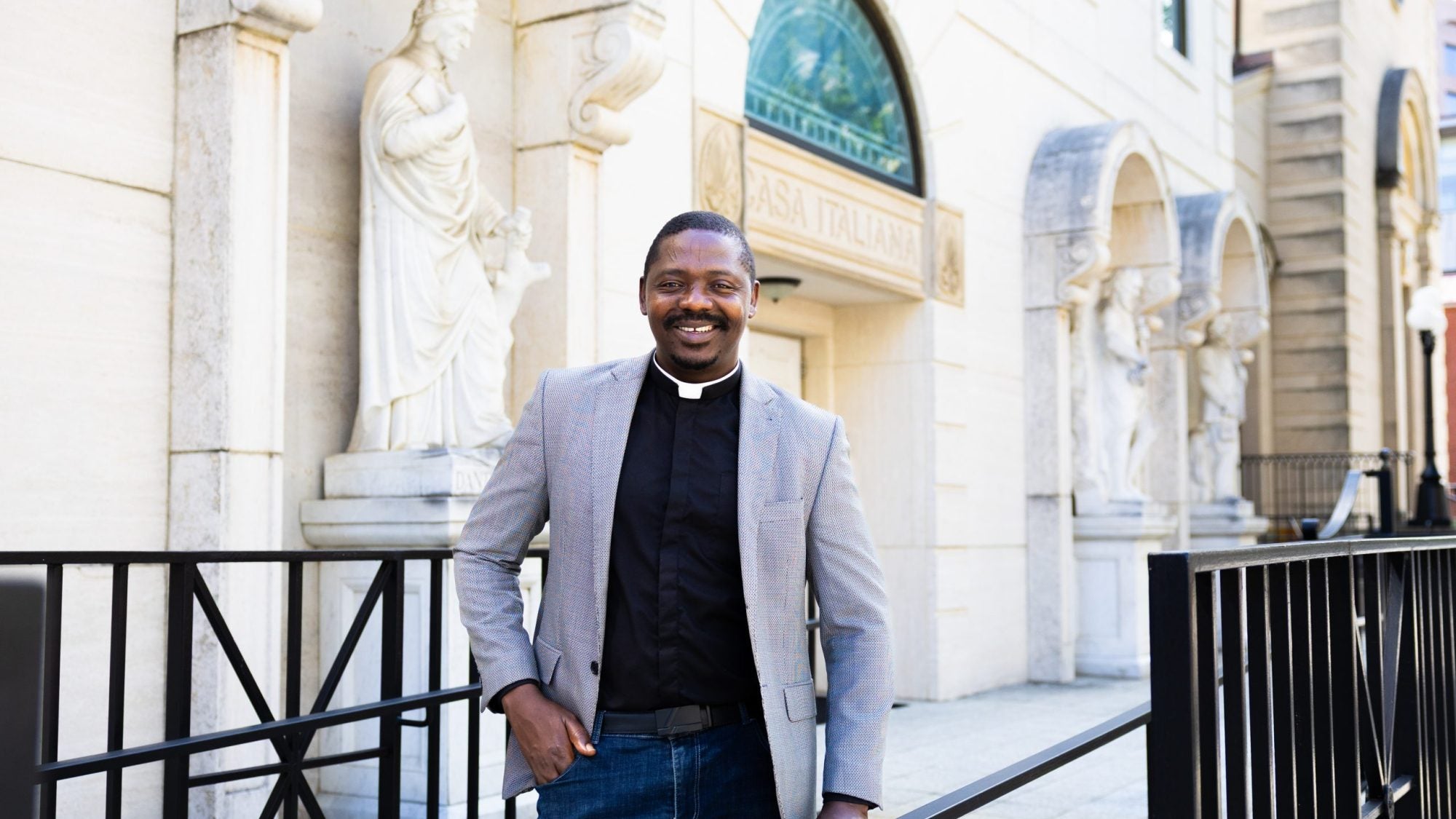 A man wearing a priest&#039;s collar and gray blazer smiles in front of a church