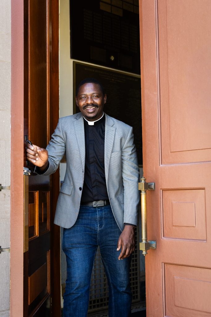 A man wearing a clerical collar opens the door of a church and smiles