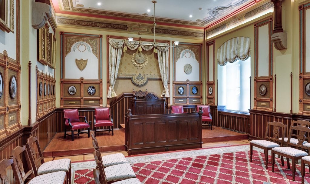 The Rostrum, a slightly elevated stage area featuring an ornate wide wooden podium. Behind it are red leather chairs. The walls are decorated with photos, wood-paneling, and curtains.