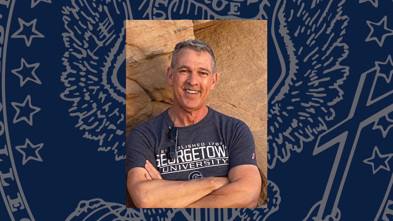 A man wears a Georgetown T-shirt and smiles with his arms crossed
