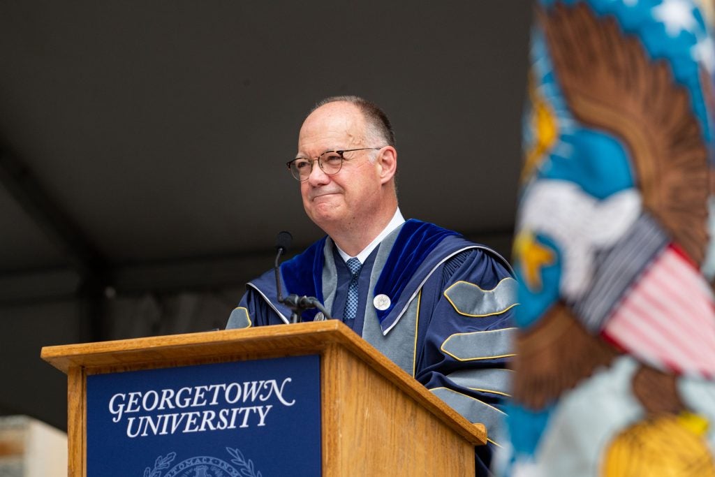 President DeGioia smiles from behind a podium wearing an academic robe