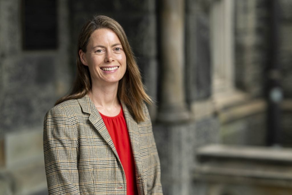 A female professor smiles in a red shirt and a plaid blazer
