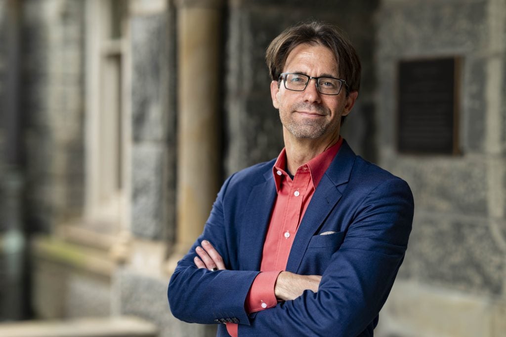 A male professor wearing glasses crosses his arms in a headshot