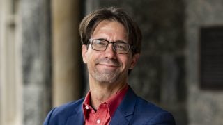 A professor wearing glasses smiles with his arms crossed