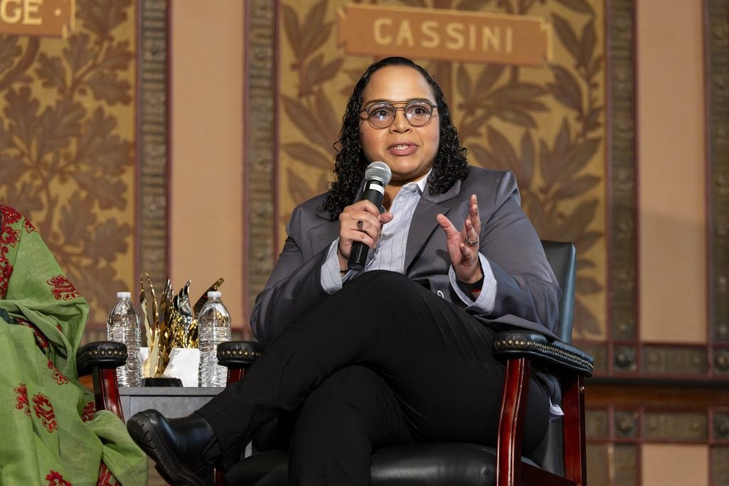 A woman in a gray suit and glasses speaks with a microphone on stage