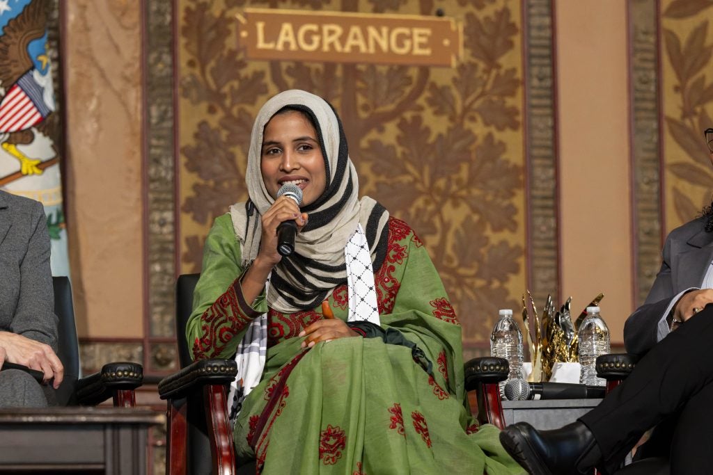 A woman in a green traditional outfit speaks with a mic on stage