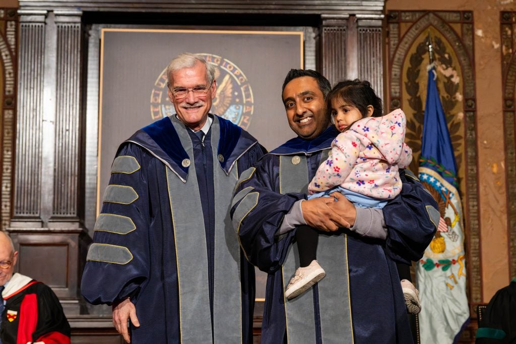A man in an academic robe holds his daughter next to the Interim President of Georgetown on a stage