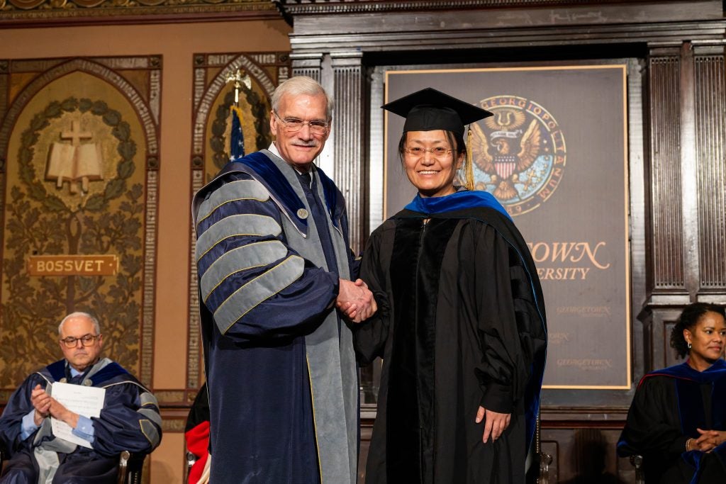 A woman in a black academic robe poses with Georgetown's interim president on a stage.