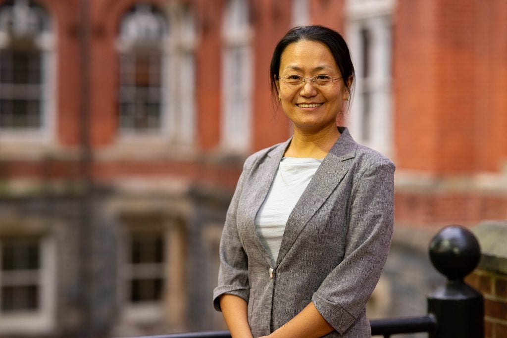 A woman in a gray blazer and glasses smiles in front of a building.