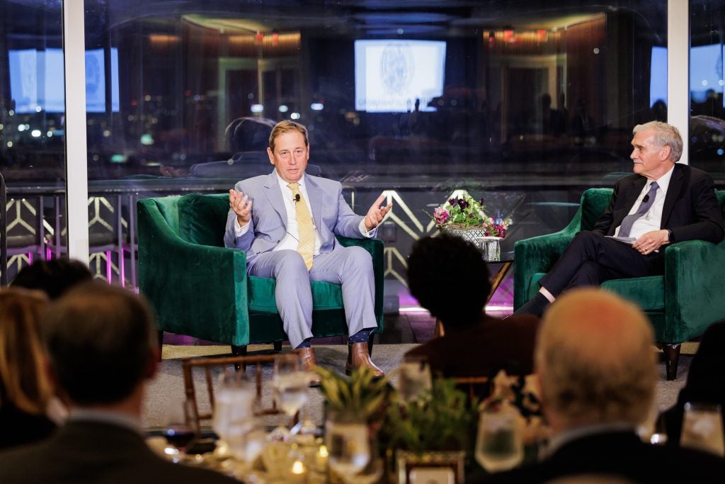 Two men sit on a stage in suits and talk in front of an audience