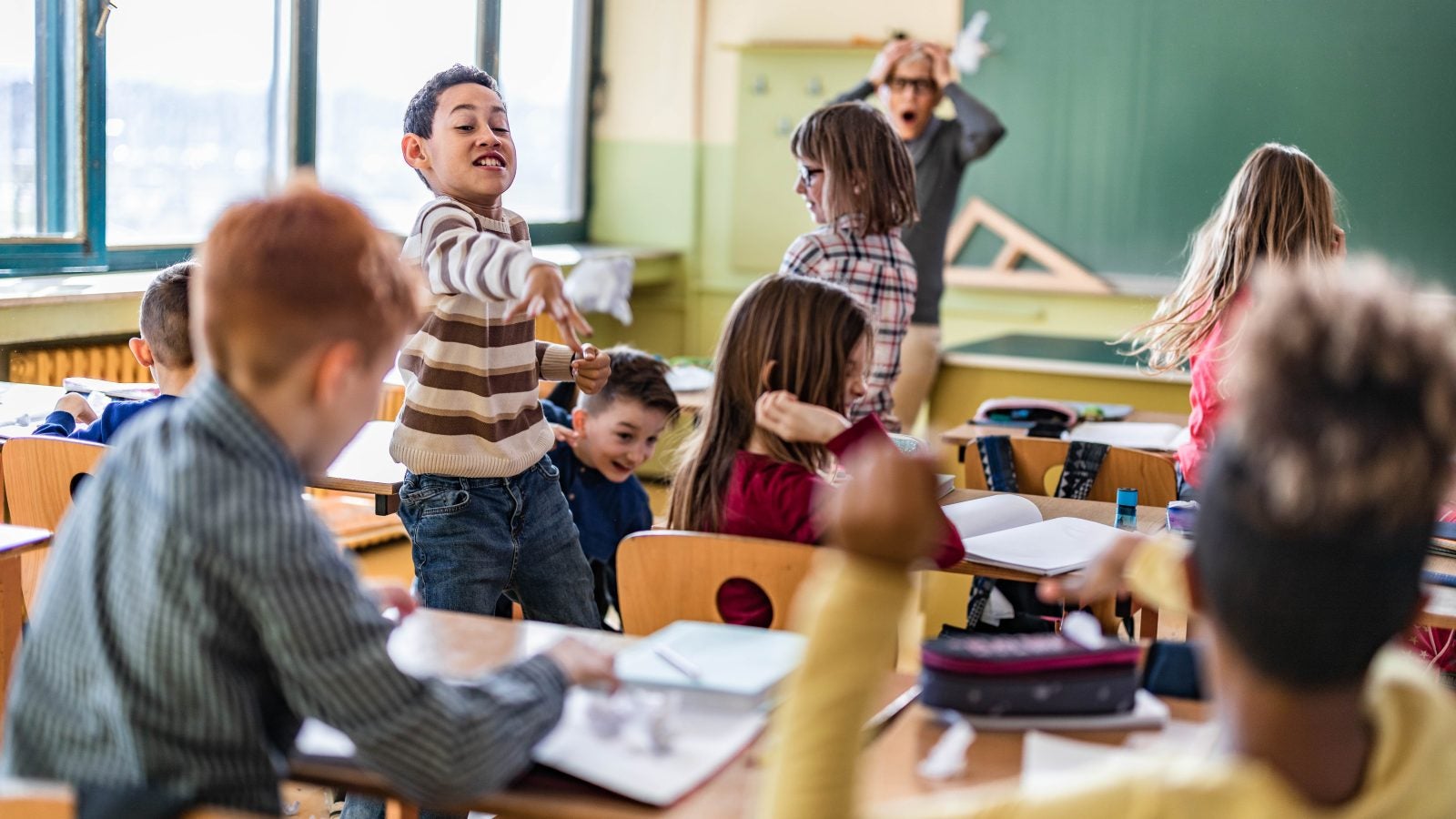 Large group of school kids making chaos during a class in the classroom while their teacher is frustrated about that.