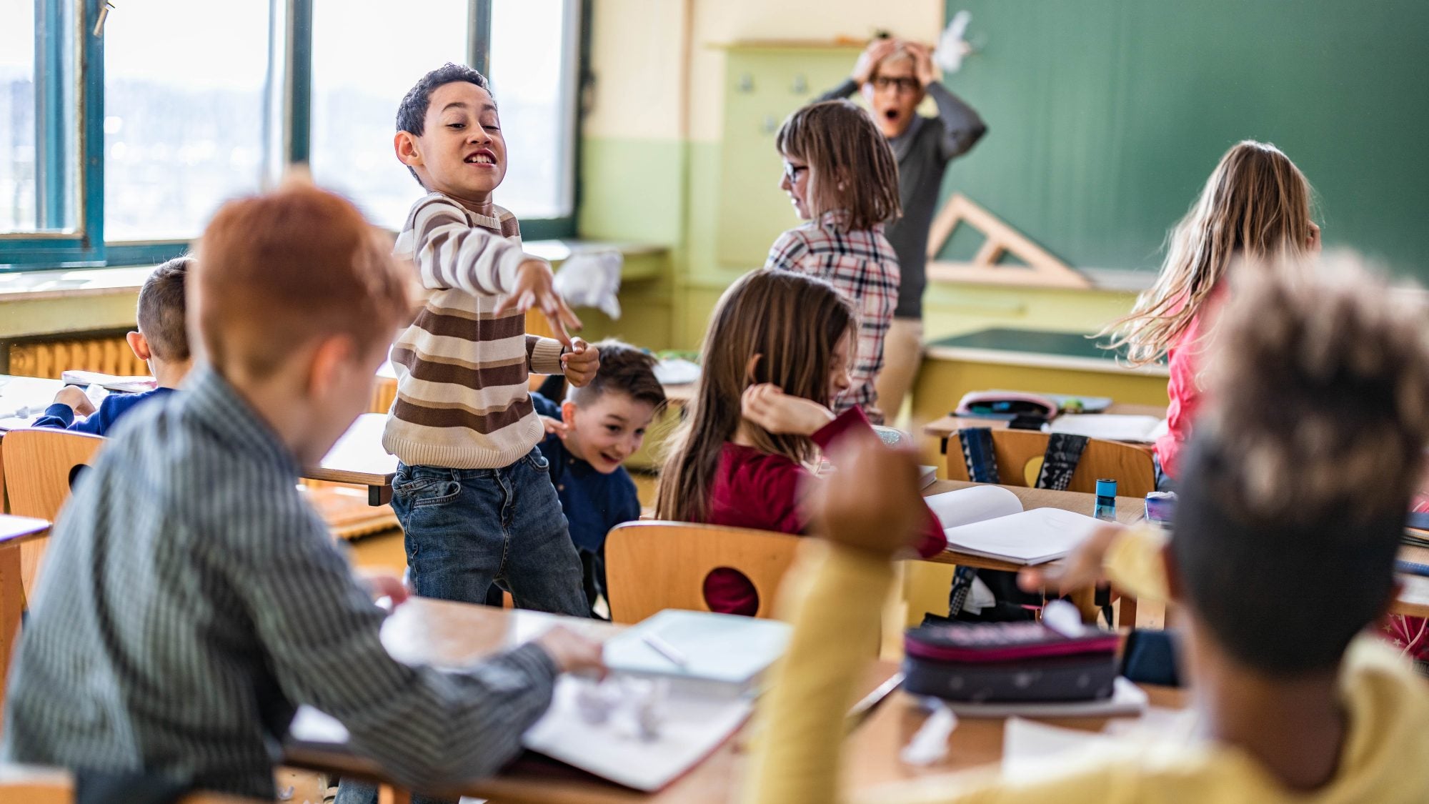 Large group of school kids making chaos during a class in the classroom while their teacher is frustrated about that.