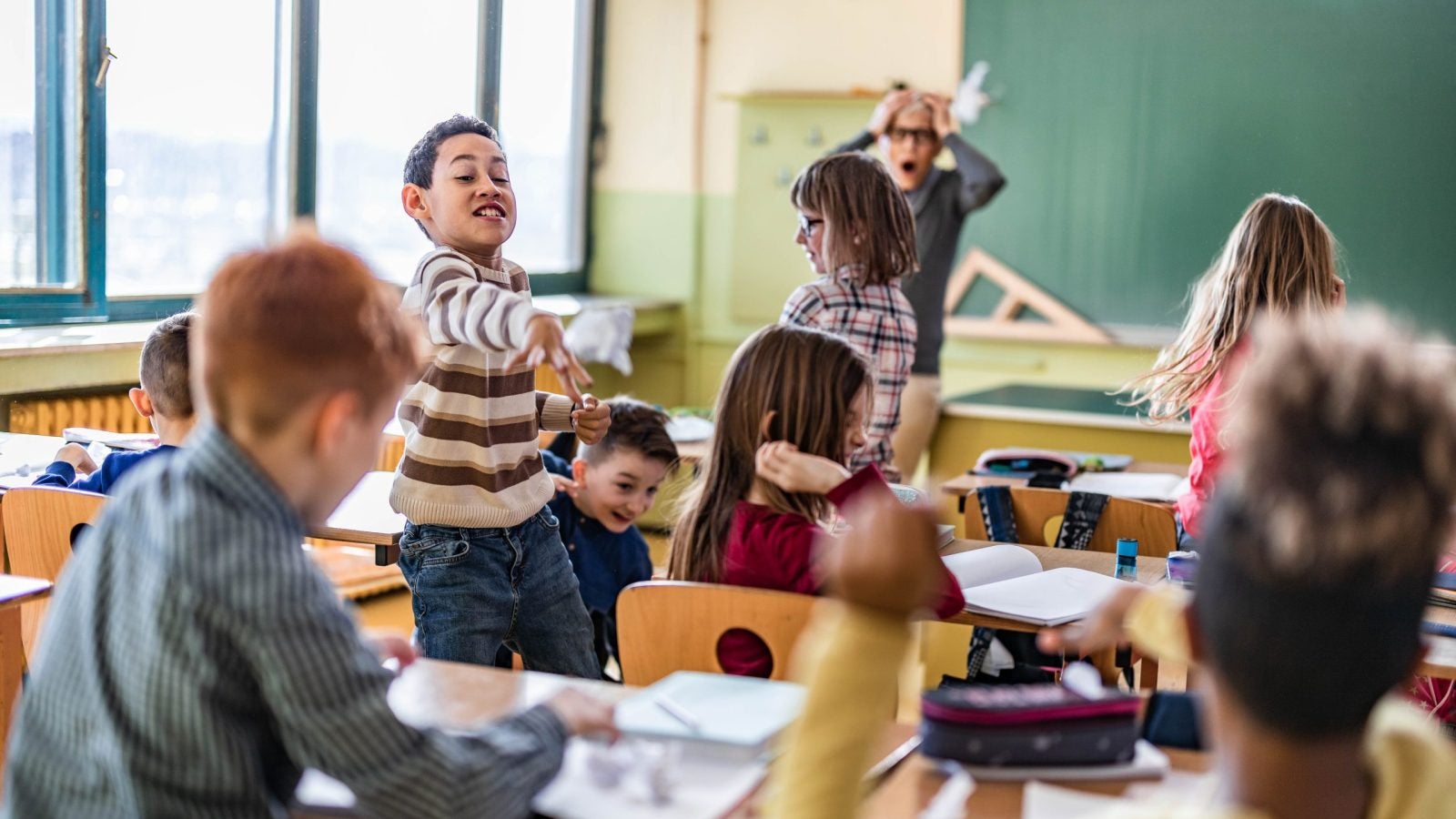Large group of school kids making chaos during a class in the classroom while their teacher is frustrated about that.