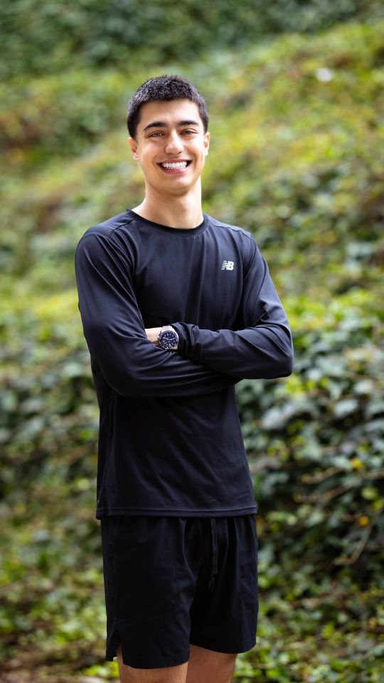 Kiumars in a black shirt with his arms crossed in front of some bushes