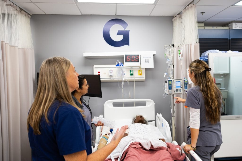 An instructor talks with student nurses gathered around the bedside of a mannequin patient. A "G" for Georgetown appears above the bed