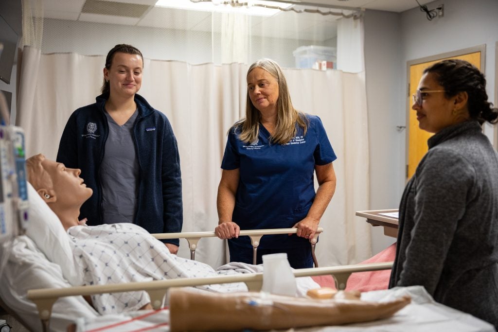 A nurse looks over a mannequin in a hospital bed as two student nurses look on