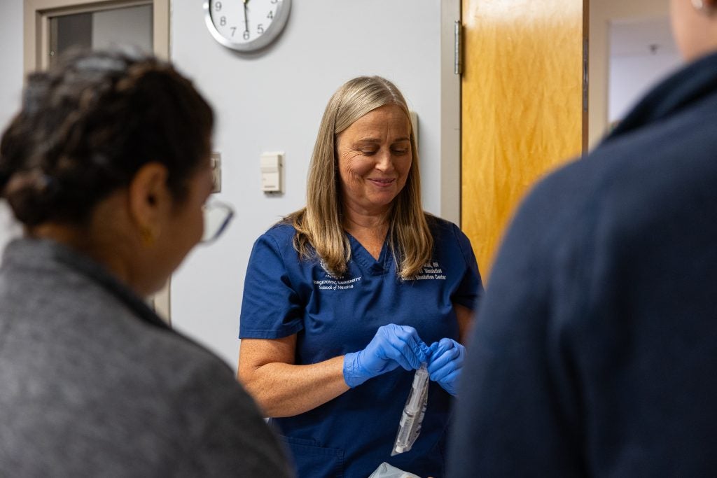An instructor smiles while opening a plastic pack of tools in front of nursing students