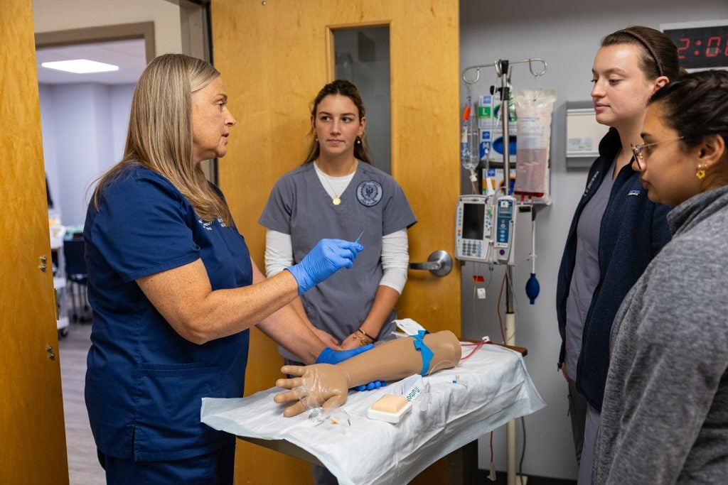 An instructor wearing blue scrubs shows student nurses how to insert an IV in a mannequin arm