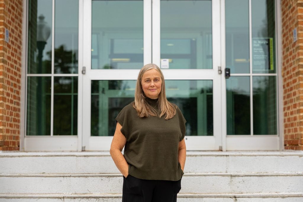 A woman in a brown shirt and black pants stands on the steps outside an academic building