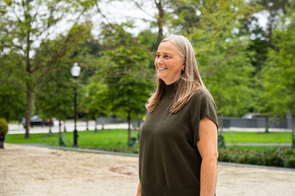A woman in a brown shirt smiles in front of a lawn and trees