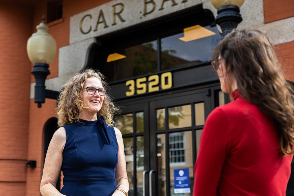 A woman with blonde curly hair smiles while talking to a student in a red sweater outside a building that says "Car Barn"