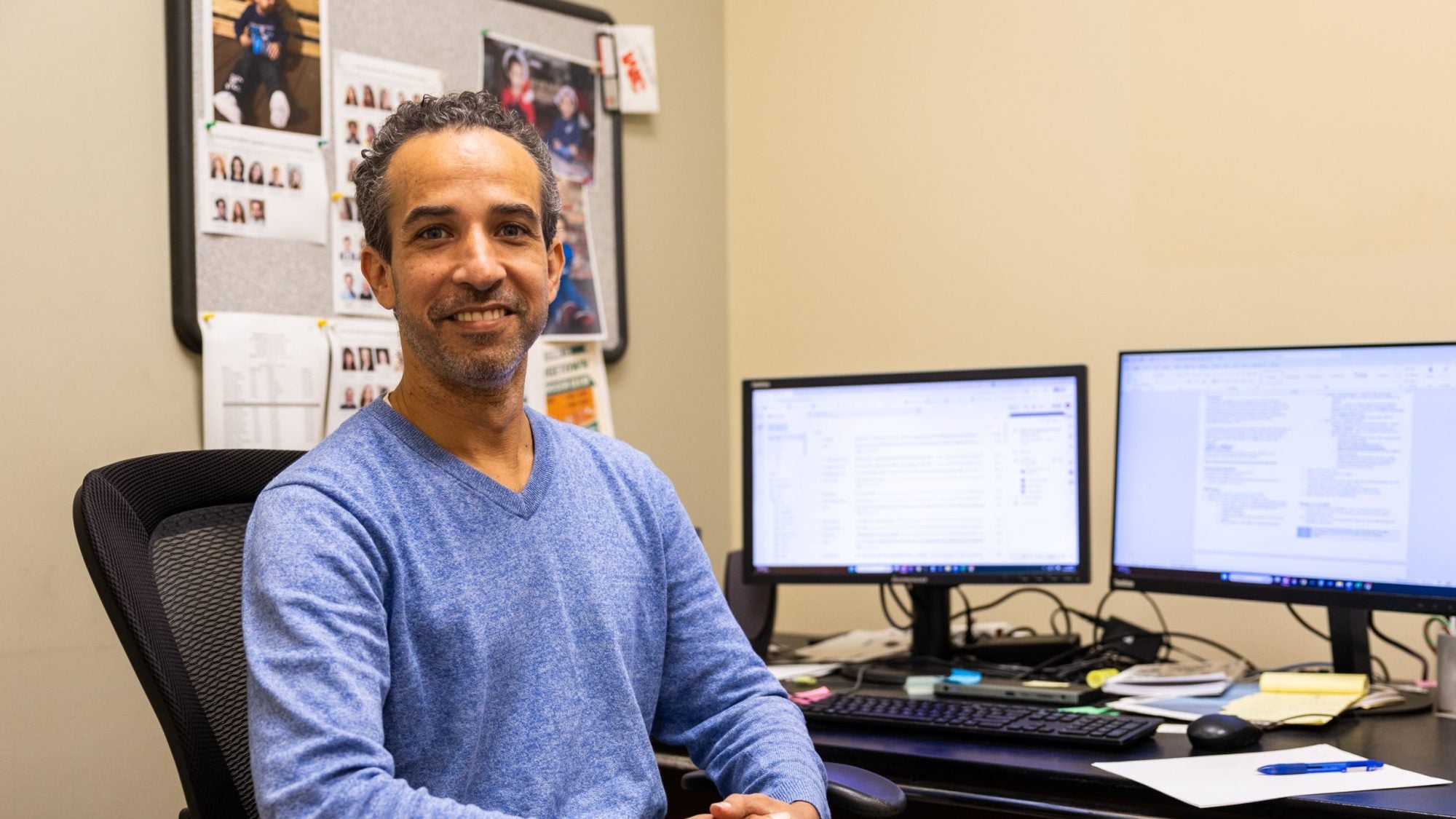 A man in a blue sweater smiles at his desk