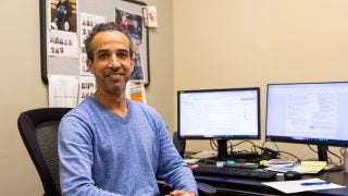 A man in a blue sweater smiles at his desk
