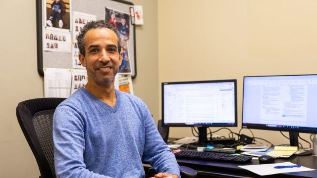 A man in a blue sweater smiles at his desk
