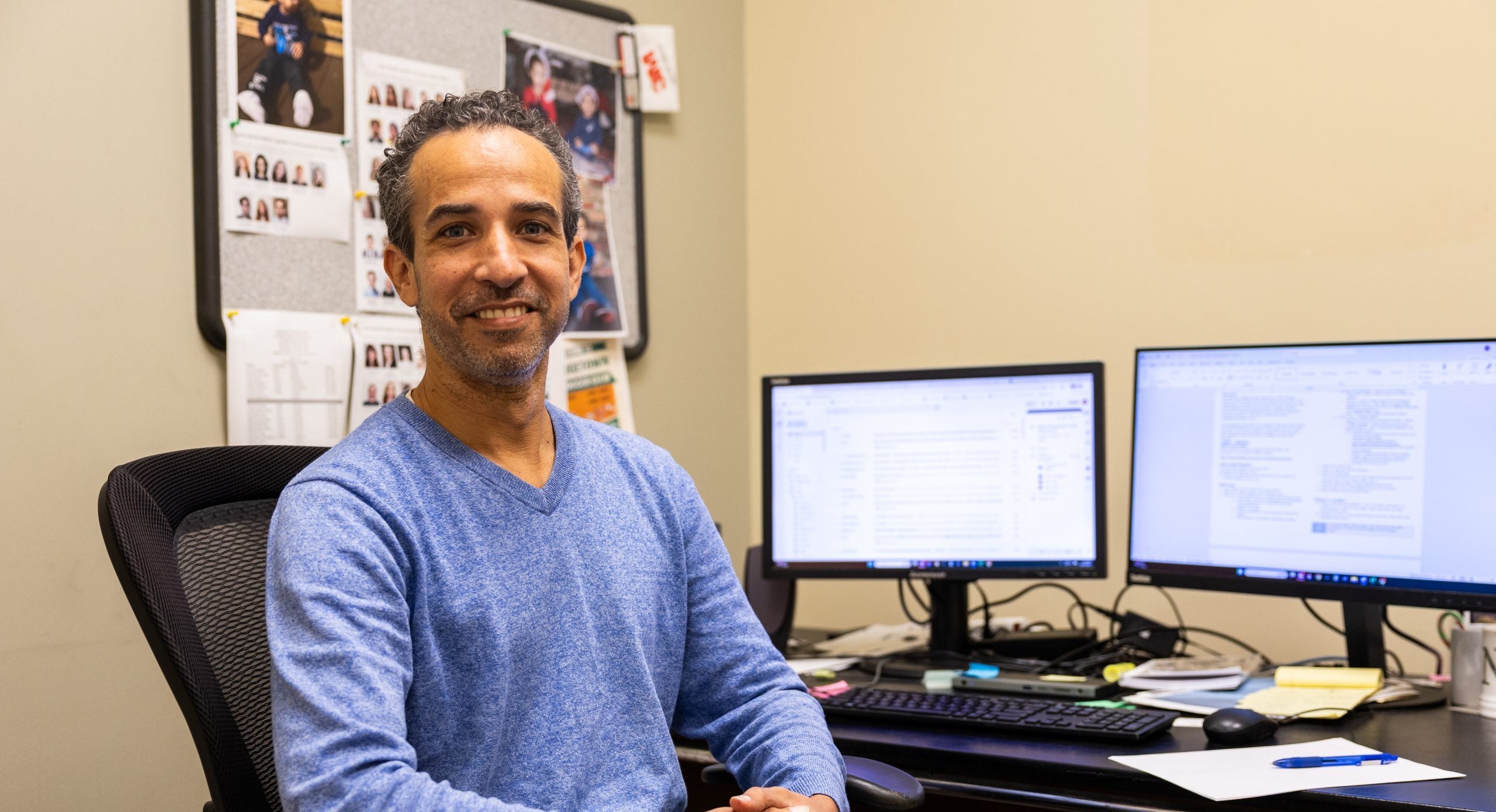 A man in a blue sweater smiles at his desk
