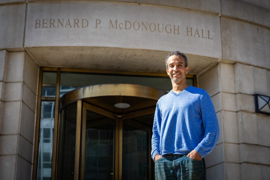 A man in a blue shirt smiles outside a building that says "McDonough Hall"
