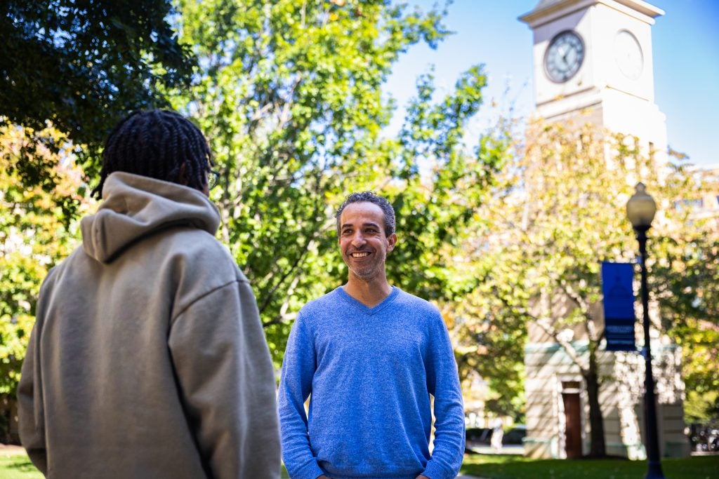 A man in a blue shirt speaks to a student outdoors on a sunny day. A clocktower is behind them