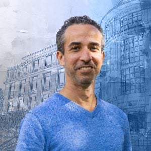 A man in a blue v-neck sweater smiles in front of a textured blue background of a building