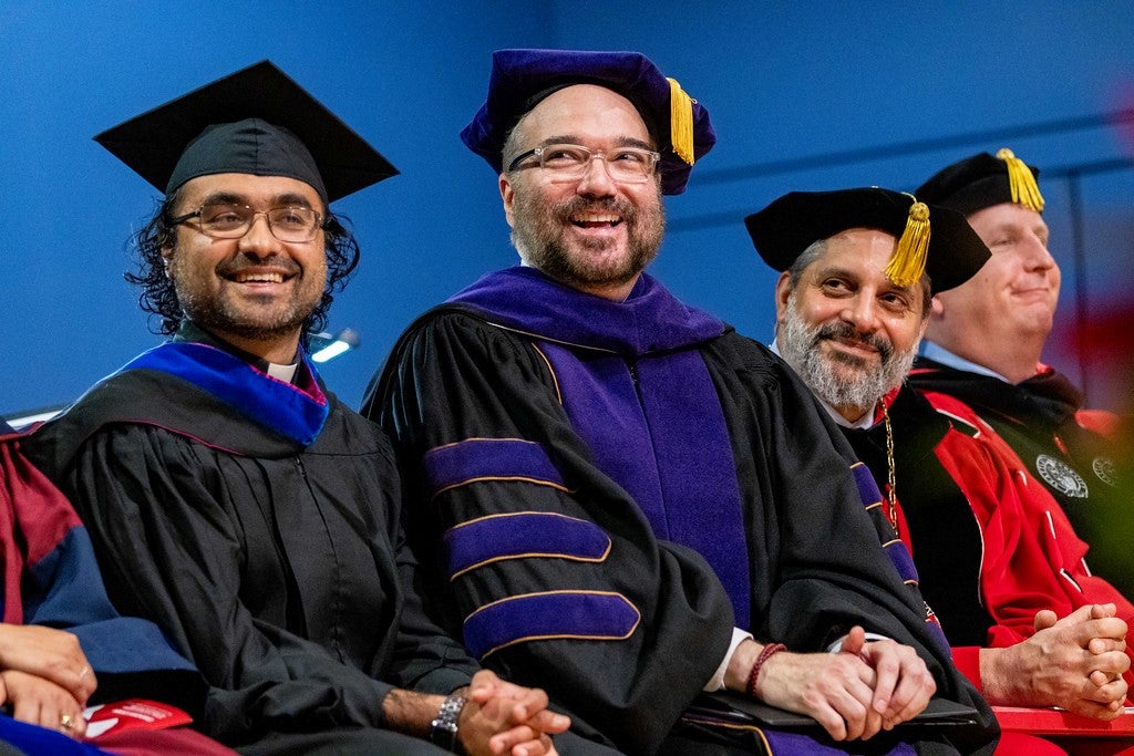 Three men in academic robes smile during a commencement