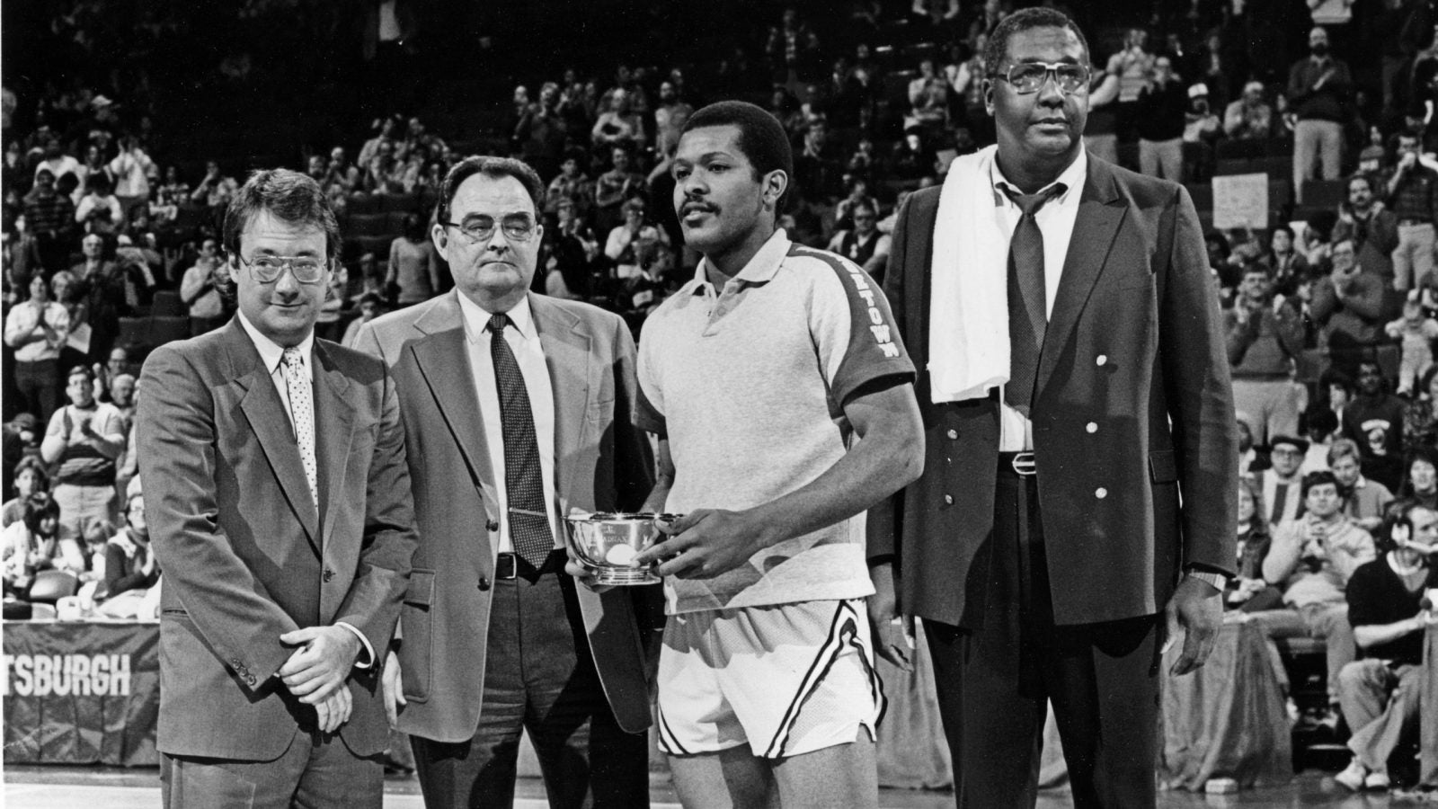 A black and white photo of President John J. DeGioia standing on a basketball court with a player holding a tropy and the late Coach John Thompson Jr.