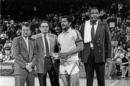 A black and white photo of President John J. DeGioia standing on a basketball court with a player holding a tropy and the late Coach John Thompson Jr.