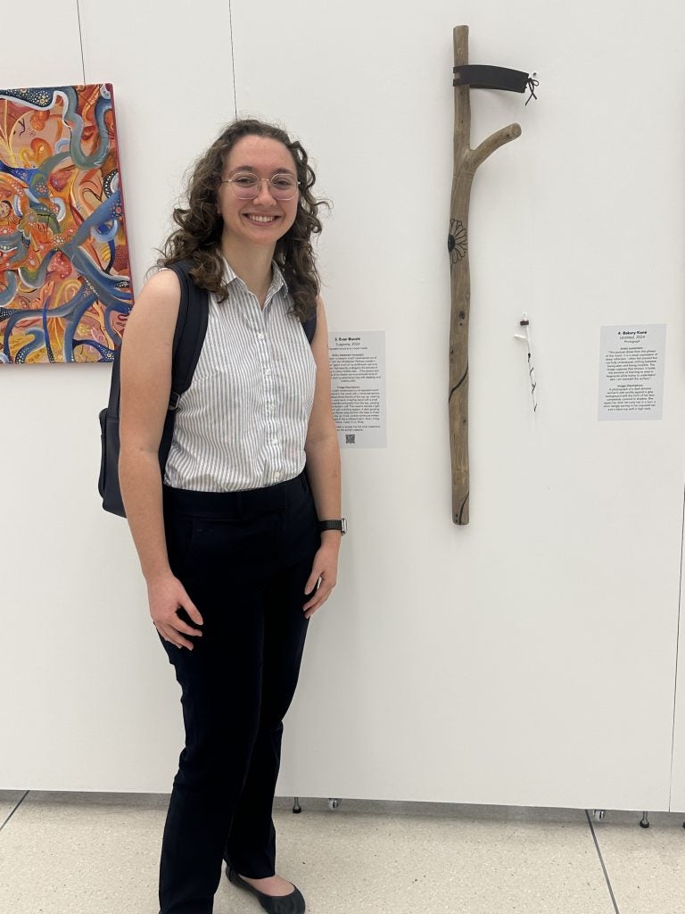 A student poses next to the mobility device she carved that hands on the wall in an exhibit