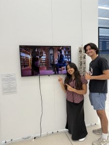 Two students smile next to a screen displayed on a white wall in an exhibit