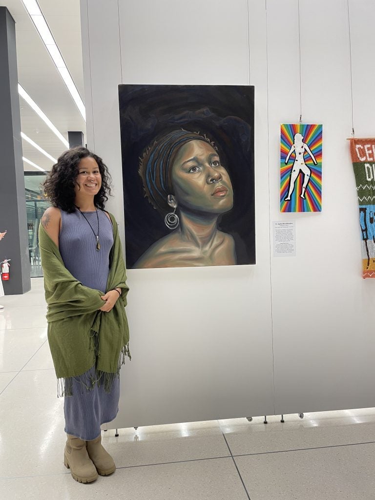 A student poses with her painting that's displayed on a white wall in an exhibit