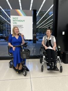 One woman in a wheelchair speaks next to another person in a wheelchair in front of windows and a sign that says "Not Without Us"