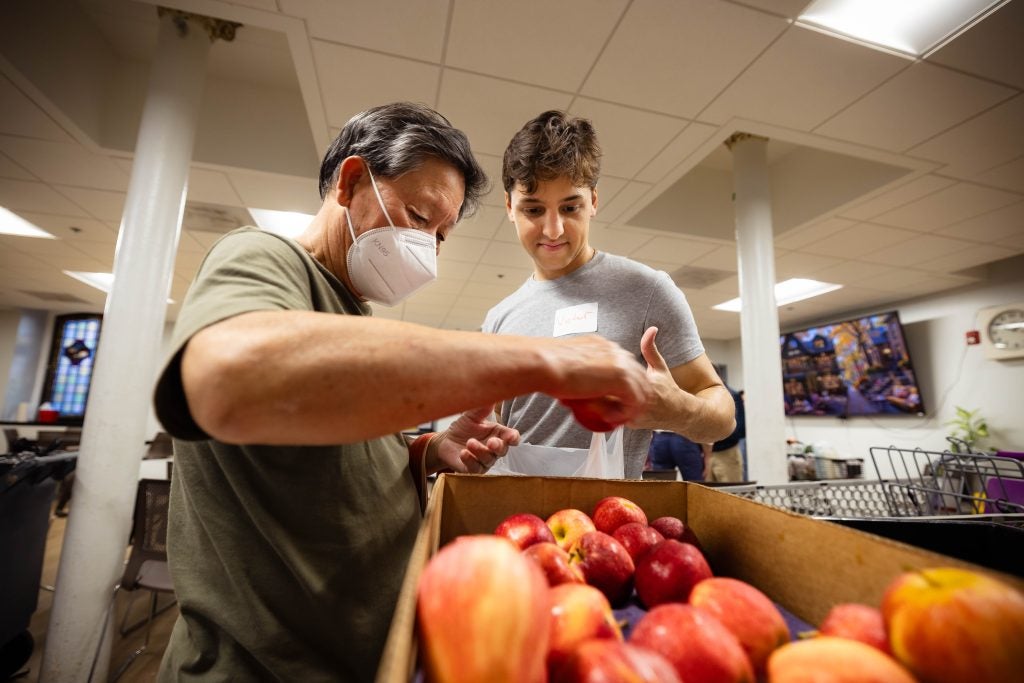 A student helps a client visiting a food pantry with groceries. A bin of apples is in front of them