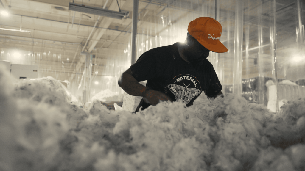 A man works with clothing waste in a factory. He wears a black shirt and an orange cap