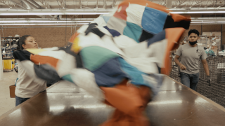 Two people lift up a brightly colored quilt off a table