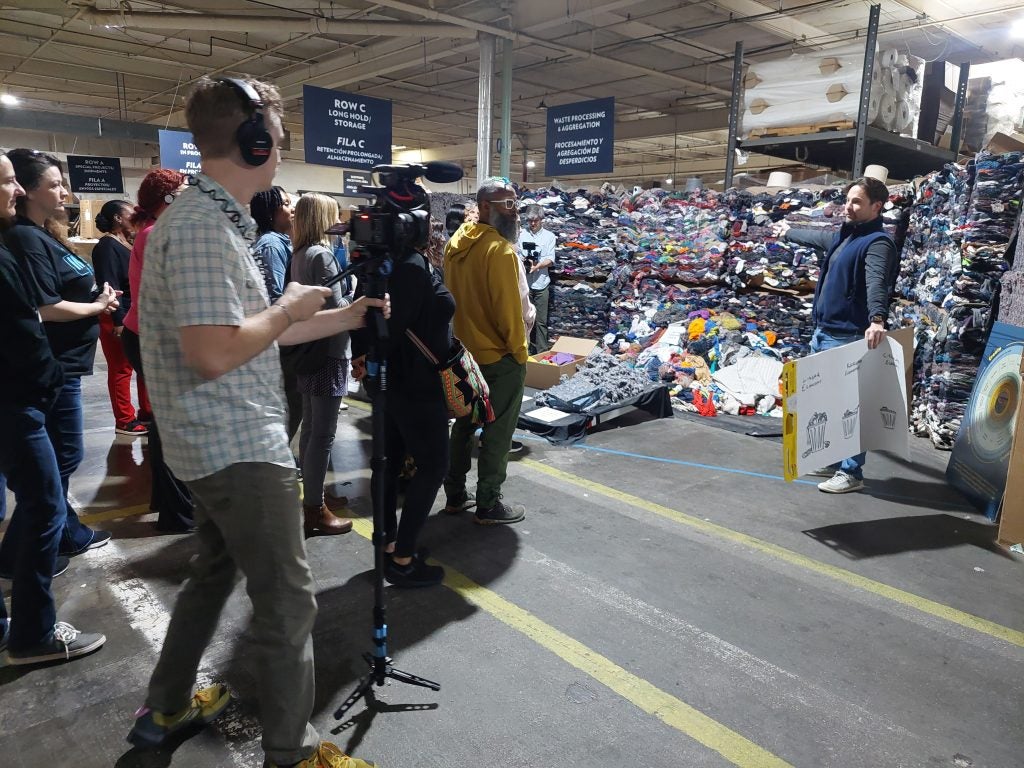 A film crew films a woman standing in front of a pile of clothing waste.