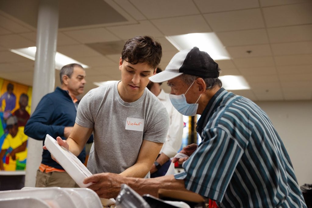 A student helps an elderly man wearing a mask and a hat with his groceries